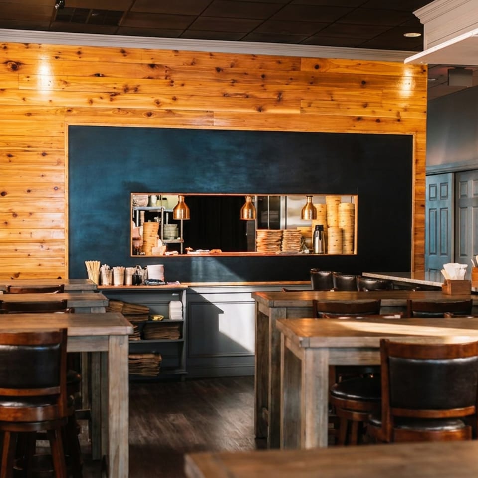 Warm, rustic restaurant dining room with wooden tables, leather chairs, a wood-paneled wall, open kitchen, and stocked bar.