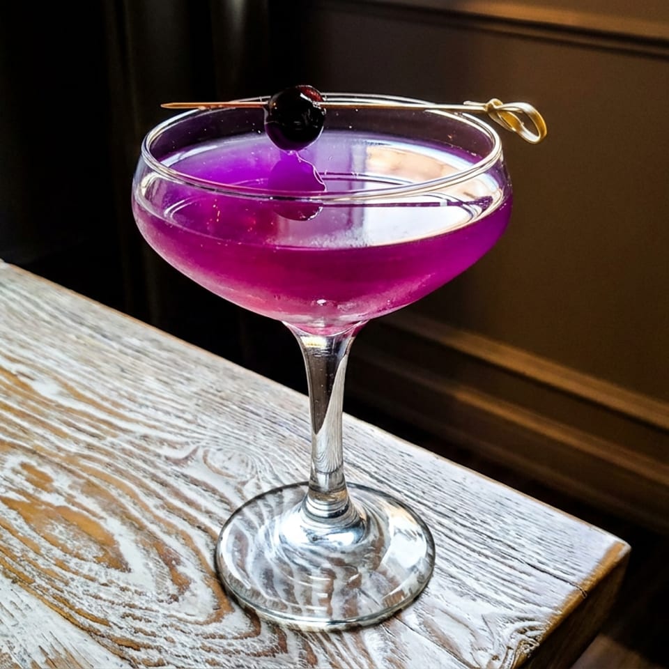 Vibrant purple cocktail in a coupe glass, garnished with a dark cherry, sits on a rustic whitewashed wooden table.